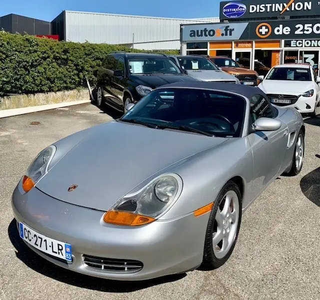 Porsche Boxster 986 gris 3/4 avant droit, capote noire, feux avant ovales et jantes argentées visibles.