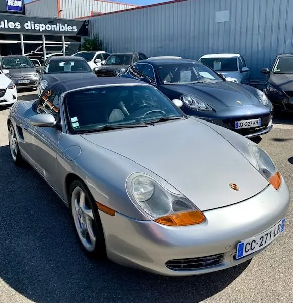Porsche Boxster 986 gris vue 3/4 avant droit, capote noire et feux avant caractéristiques orange, stationnée en extérieur.
