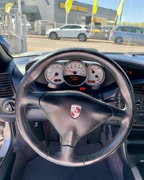 Vue intérieure centrée sur le volant noir avec logo Porsche et tableau de bord blanc d'une Boxster 986 gris 2001.