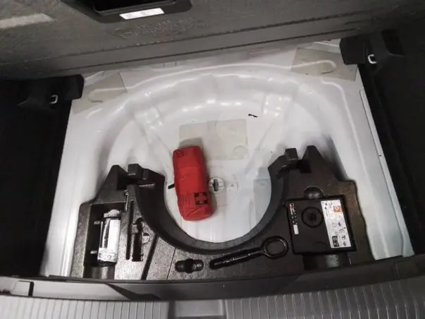 Vue plongeante sur le coffre ouvert d’un Volkswagen T-Roc blanc, montrant le rangement sous la moquette avec kit de secours rouge.