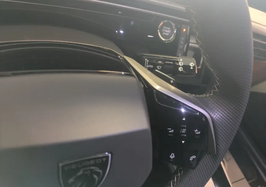 Gros plan sur le volant perforé et les commandes tactiles du Peugeot 5008 Hybride gris titane, avec bouton start/stop visible.