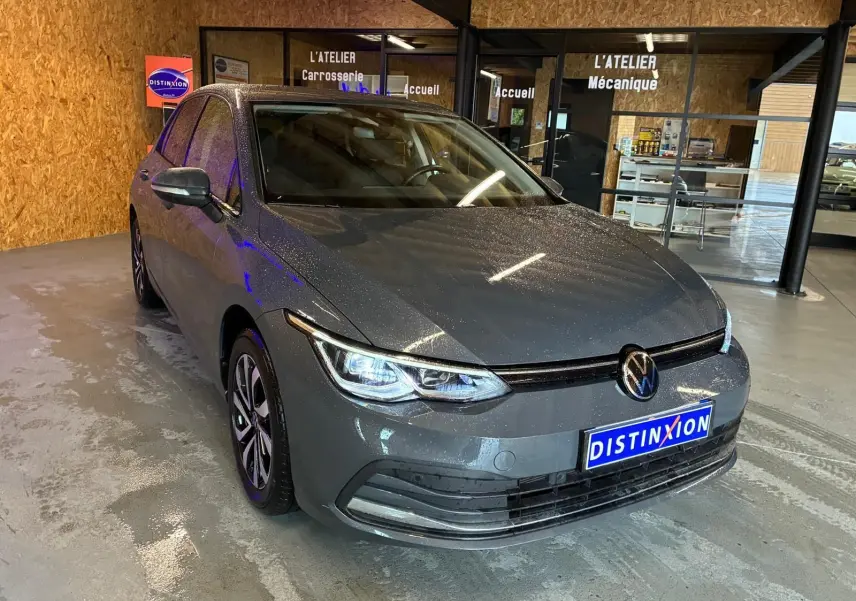 Vue 3/4 avant d'une Volkswagen Golf gris métallisé 2022 avec phares allumés dans un atelier automobile.