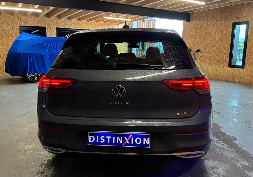 Vue arrière d'une Volkswagen Golf gris foncé 1.5 eTSI 130 DSG7 ACTIVE avec feux arrière allumés en intérieur.