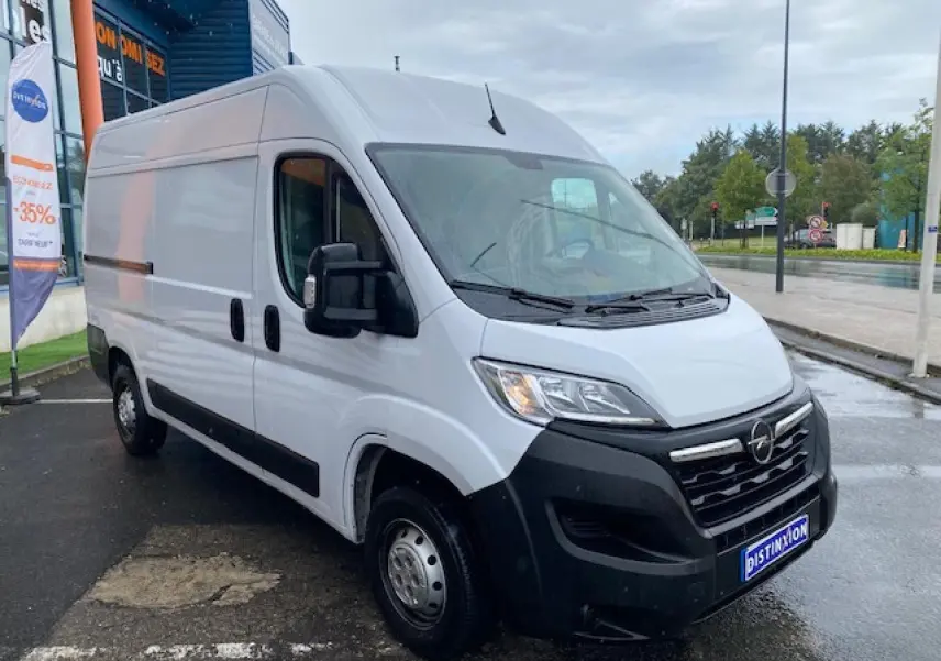 Opel Movano Fourgon blanc vu en 3/4 avant droit sur parking humide devant un bâtiment commercial.