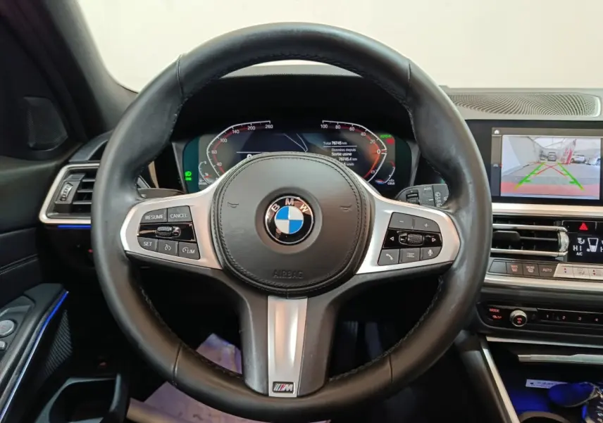Vue intérieure centrée sur le volant cuir noir BMW avec tableau de bord digital et écran multimédia en arrière-plan.