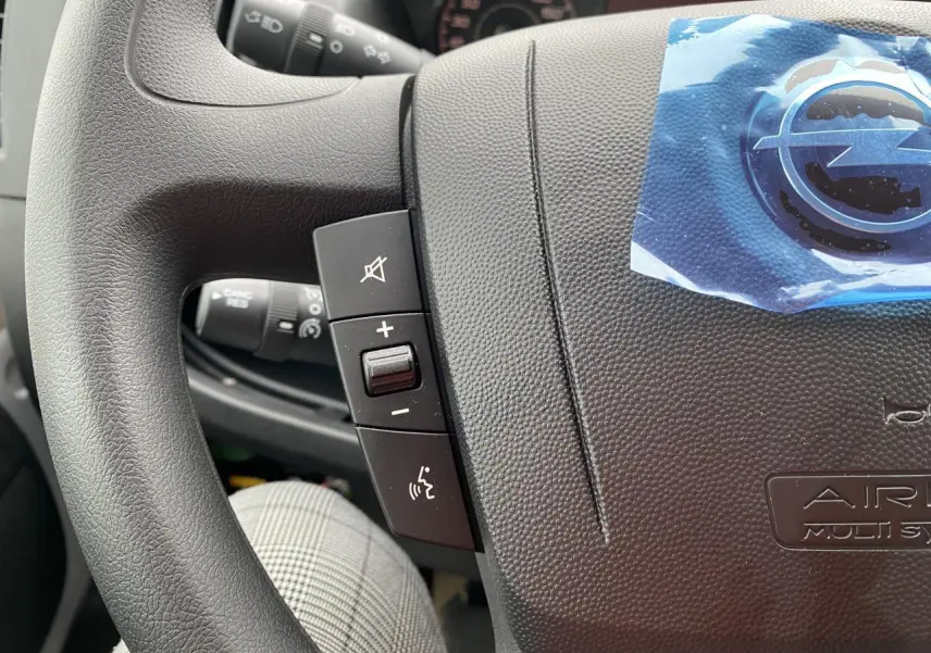Gros plan sur le volant noir de l'Opel Movano Fourgon 2022 avec commandes audio et logo protégé par un film bleu.