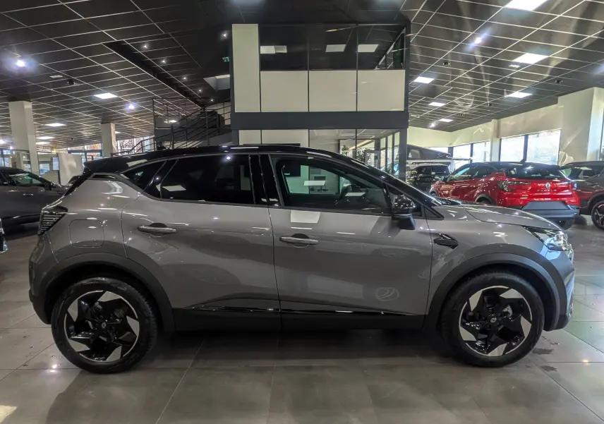 Profil côté gauche du Renault Captur 2025 gris Cassiopé avec toit noir et jantes biton dans un showroom.
