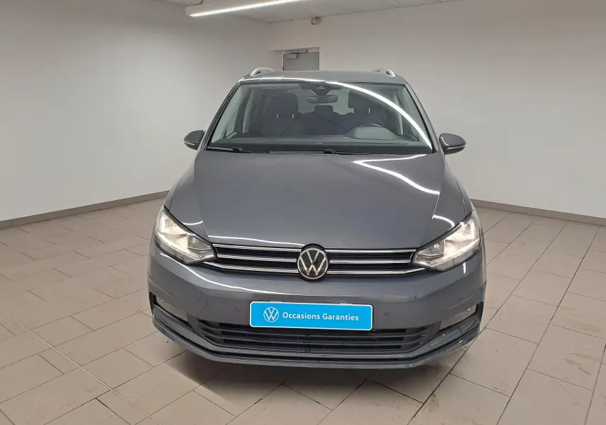 Vue avant d'un Volkswagen Touran gris dauphin 2024 avec phares allumés dans un showroom lumineux.