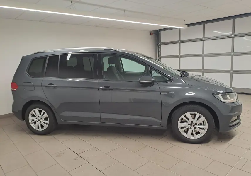 Vue de profil côté gauche d'un Volkswagen Touran 2024 gris dauphin, stationné en intérieur avec jantes alliage visibles.