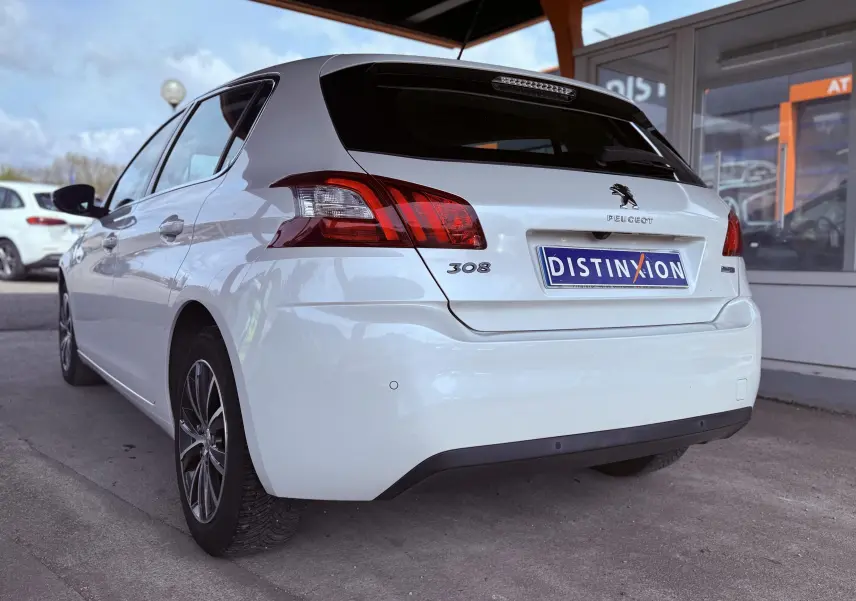 Vue 3/4 arrière droite d'une Peugeot 308 blanche nacrée avec feux arrière distinctifs et logo Peugeot visible.