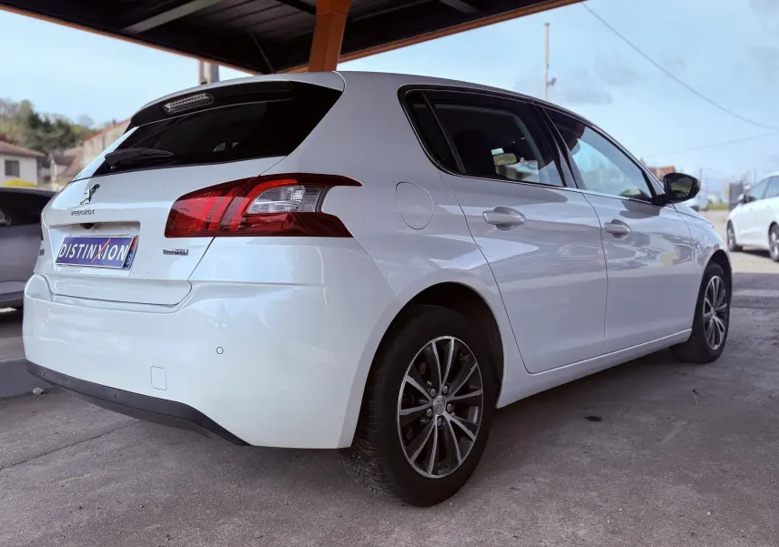 Peugeot 308 blanc nacré vue 3/4 arrière droit sous abri, avec feux arrière rouges et jantes alliage foncé.
