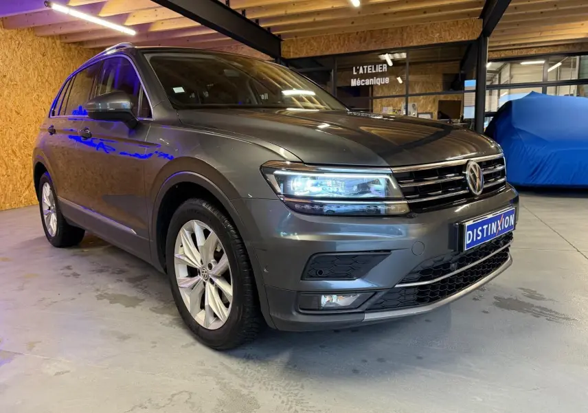 Volkswagen Tiguan gris 3/4 avant droit, phares allumés, jantes alu, dans un garage intérieur.