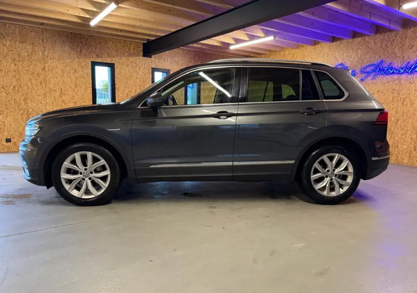 Vue de profil côté gauche d'un Volkswagen Tiguan gris avec jantes alu et barres de toit dans un garage.