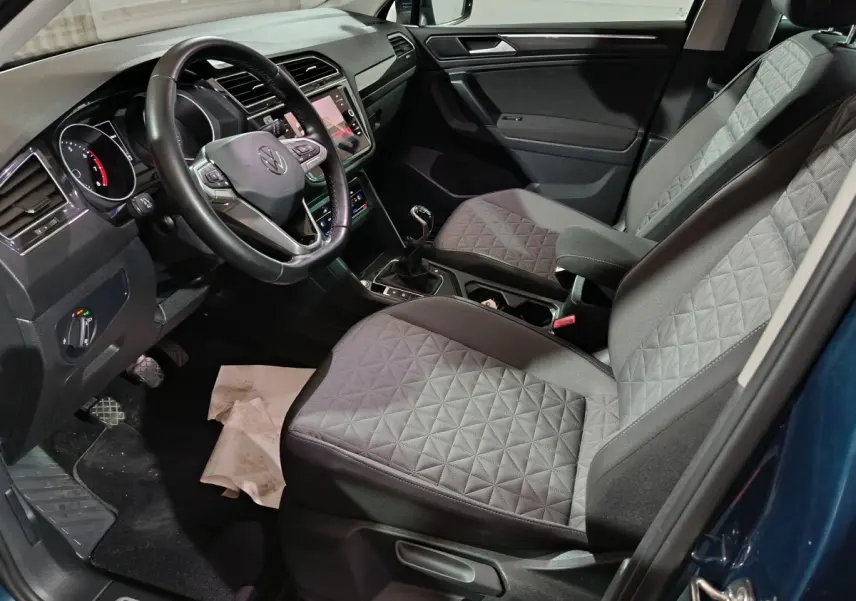 Vue intérieure côté conducteur du Volkswagen Tiguan bleu, montrant le volant cuir multifonctions et les sièges avant gris à motifs.