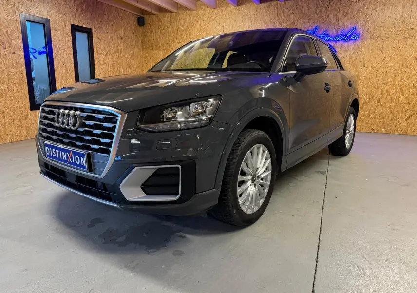 Audi Q2 gris en 3/4 avant droit dans un intérieur avec jantes alu et calandre chromée visible.