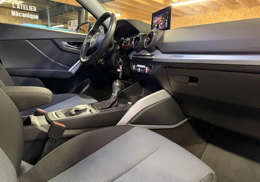 Intérieur Audi Q2 gris vu côté passager, tableau de bord avec écran GPS et console centrale avec levier de vitesse.