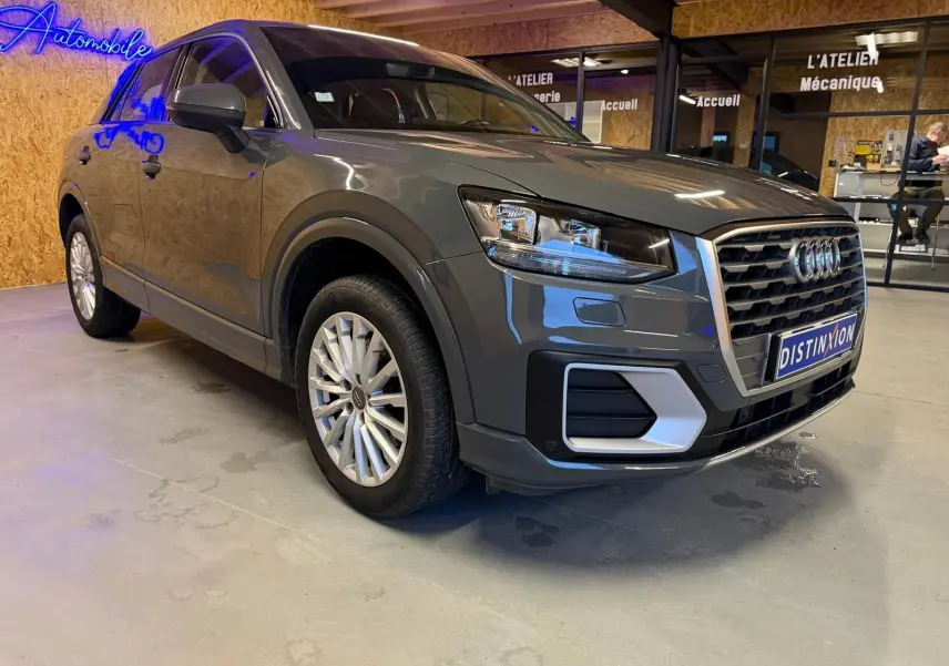 Audi Q2 gris vue 3/4 avant droit, jantes alu, calandre hexagonale et rétroviseurs électriques rabattables.