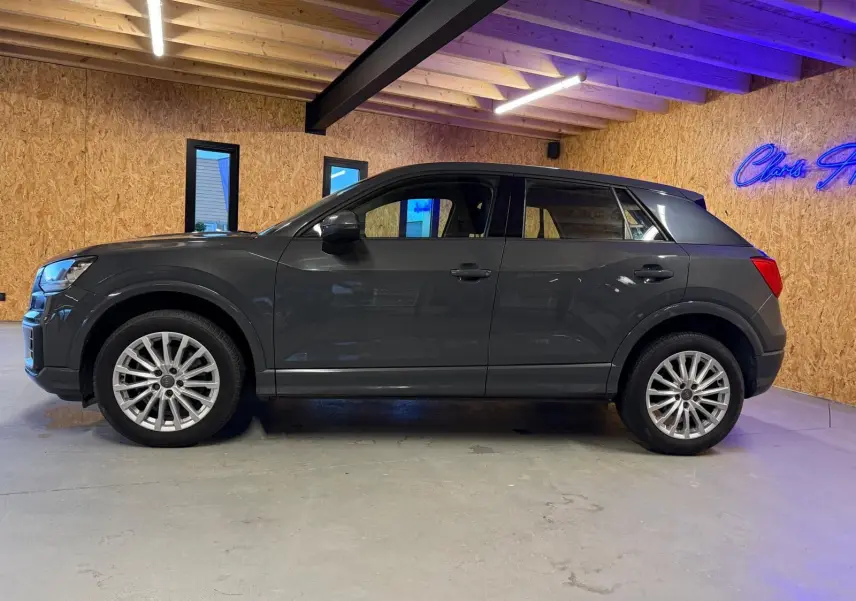 Audi Q2 gris vue de profil côté gauche dans un intérieur avec jantes alu et rétroviseurs électriques rabattables.