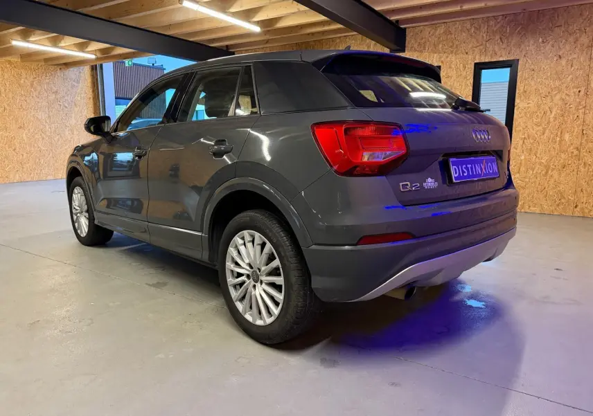 Audi Q2 gris vue 3/4 arrière droit en intérieur, avec jantes alu et sorties d'échappement chromées visibles.