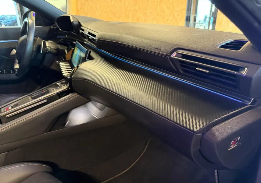 Vue intérieure côté passager du tableau de bord noir avec finition carbone et éclairage d'ambiance bleu sur Peugeot 508 SW 2024.