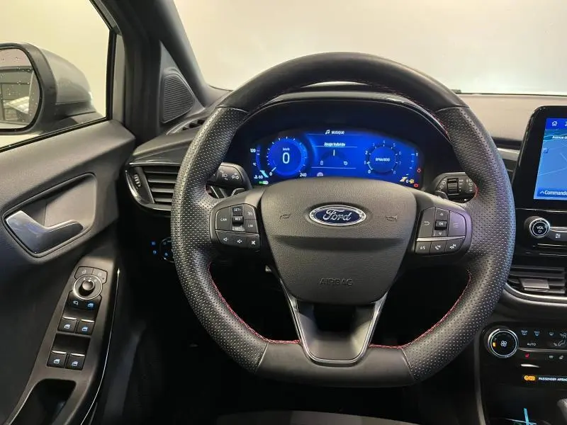 Vue centrée sur le volant sport noir avec surpiqûres rouges du Ford Puma 1.0 EcoBoost, tableau de bord numérique bleu visible