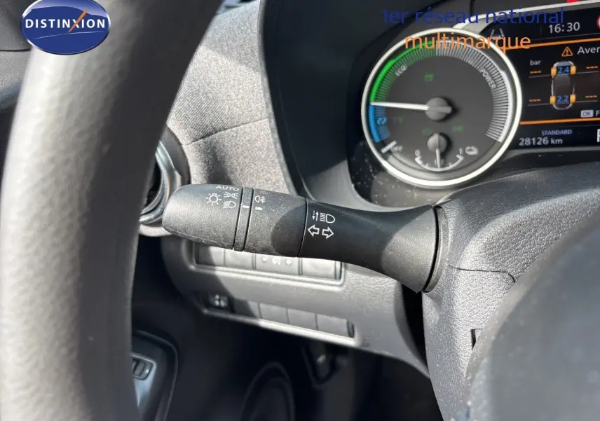 Gros plan sur le commodo d’éclairage à gauche du volant du Nissan Juke Hybrid gris, avec tableau de bord numérique en arrière-plan.