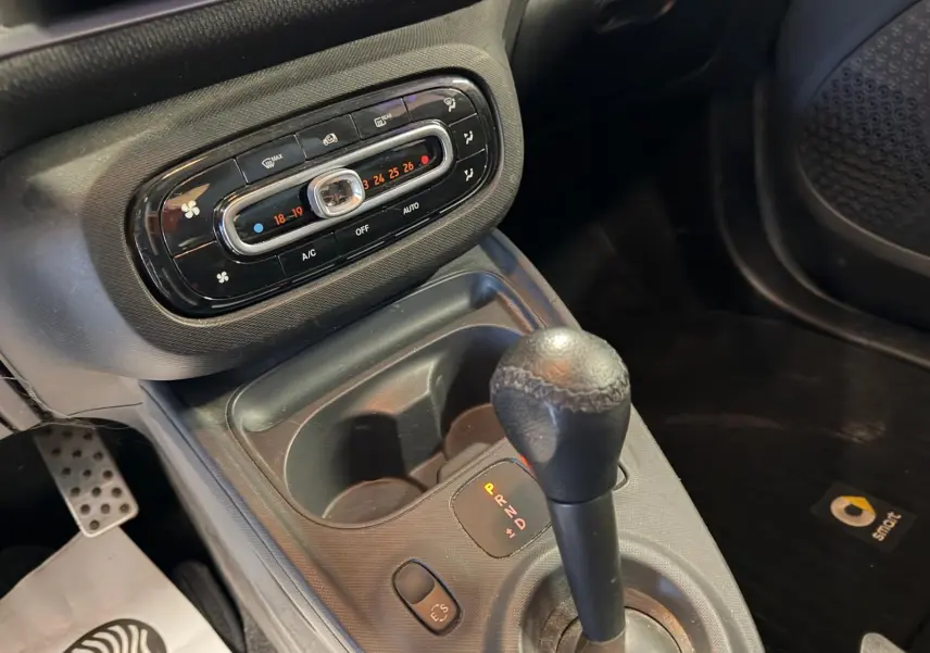 Gros plan intérieur sur la console centrale et levier de vitesse de la Smart Fortwo Cabrio noire, avec commandes climatisation et tapis logo Smart.