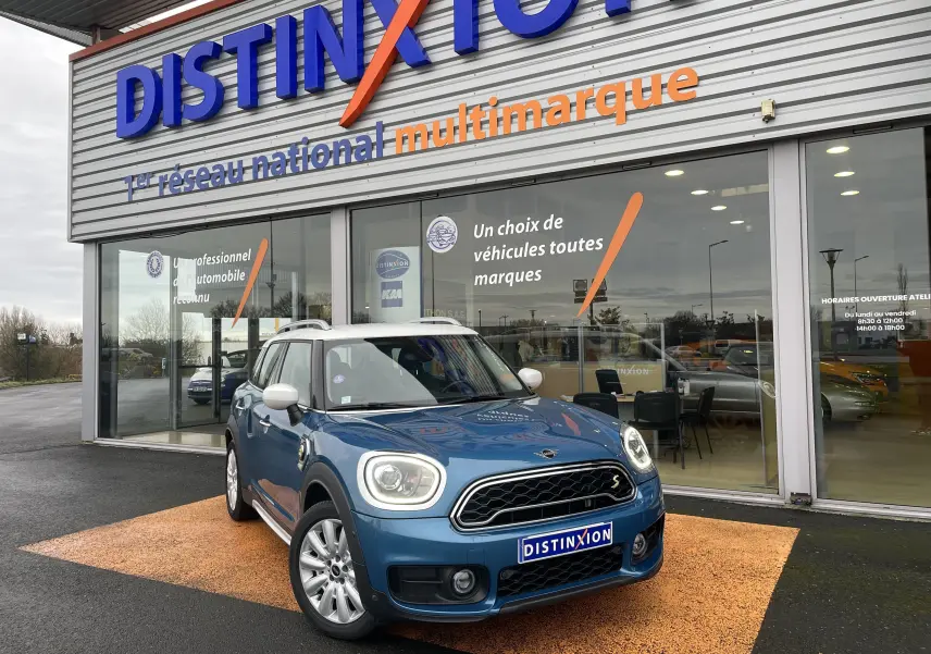 Vue 3/4 avant d'une MINI Countryman Cooper S hybride bleu Island Metal avec toit blanc devant un showroom Distinxion.