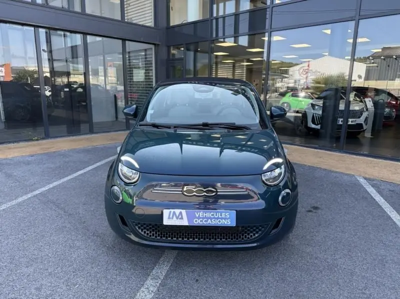 Vue de face d'une FIAT 500C 118 cv icone électrique 2021 en vert foncé métallisé, devant une vitrine de concession.