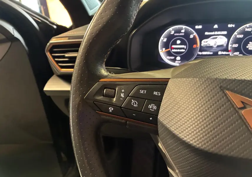 Gros plan sur le volant noir perforé avec logo CUPRA cuivré et commandes intégrées, tableau de bord numérique en arrière-plan.