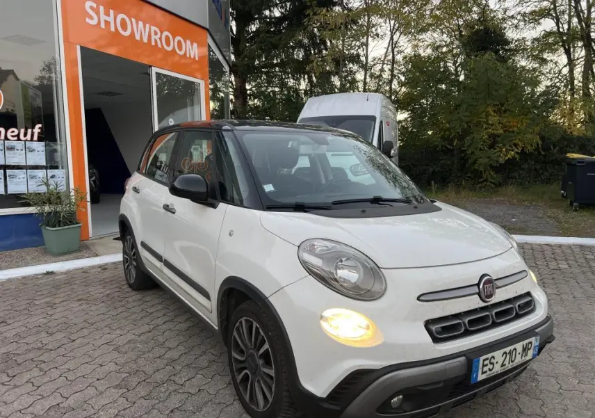 Vue 3/4 avant droite d'une Fiat 500L blanche avec toit noir bi-color, feux allumés, devant un showroom.