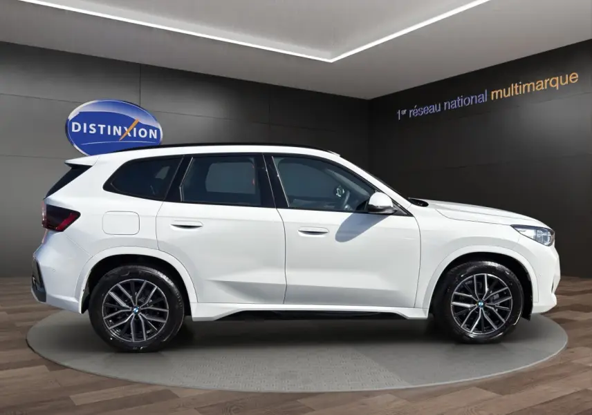 BMW X1 sDrive20d blanc vue 3/4 arrière droit, avec jantes noires et aileron de toit visible.