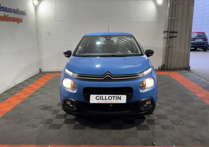 Citroën C3 bleu vue de face avec phares allumés dans un garage au sol noir et orange.
