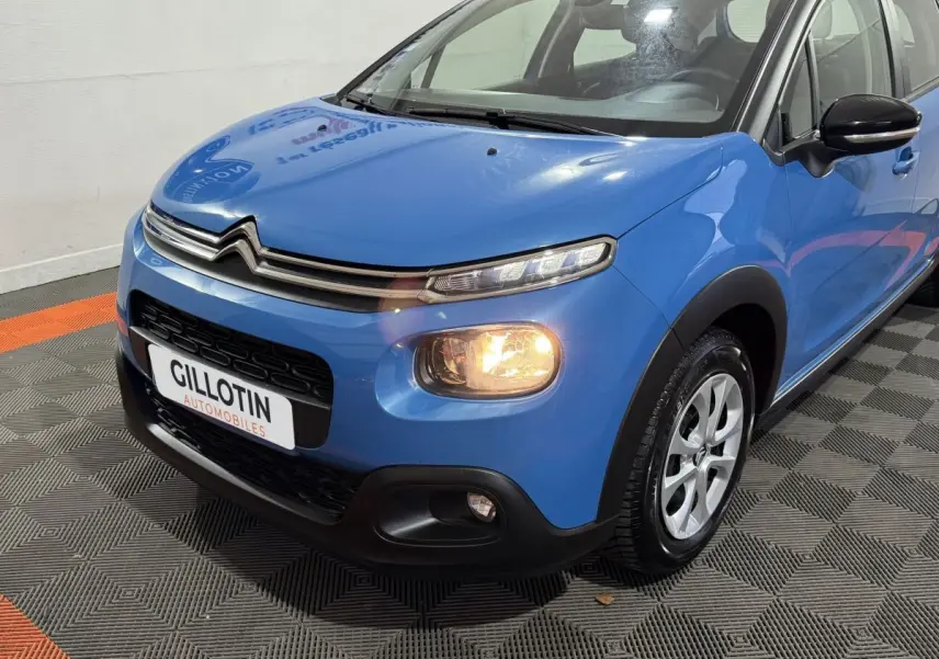 Vue 3/4 avant droit d'une Citroën C3 bleu avec feux allumés et jantes alliage visibles sur sol intérieur.