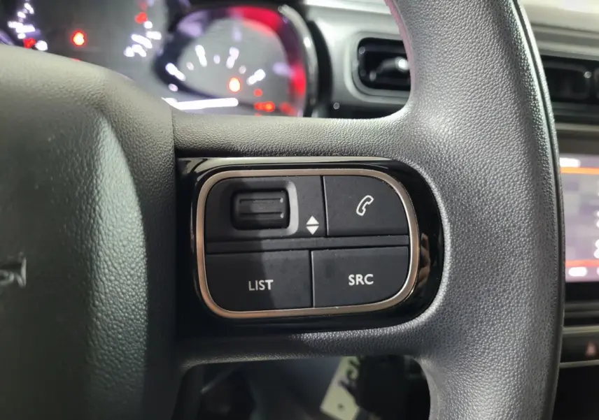 Gros plan sur les commandes gauche du volant noir de la Citroën C3 beige, avec tableau de bord et écran tactile en arrière-plan.