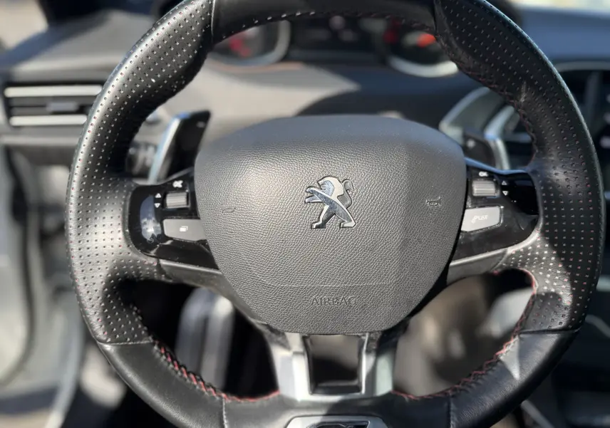 Gros plan sur le volant en cuir noir perforé avec surpiqûres rouges du Peugeot 308 SW blanc nacré, version GT.