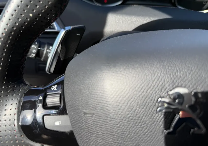 Gros plan sur le volant noir perforé avec logo Peugeot argenté et commandes intégrées, intérieur du 308 SW blanc nacré.