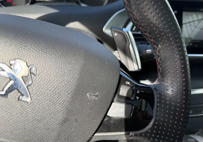Gros plan sur le volant cuir noir à surpiqûres rouges du Peugeot 308 SW blanc nacré, avec commandes intégrées et palettes au volant.