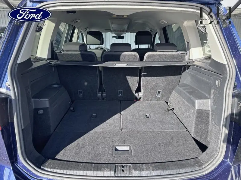 Vue arrière du coffre ouvert du Volkswagen Touran bleu Atlantique métallisé, montrant l'espace de chargement et les sièges rabattus.