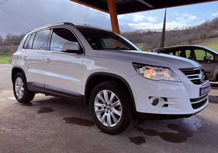 Volkswagen Tiguan blanc vu en 3/4 avant droit sous abri, avec jantes alliage et phares allumés.