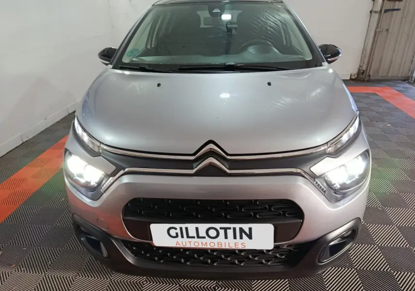 Vue avant d'une Citroën C3 gris foncé avec phares allumés dans un garage au sol carrelé noir et orange.