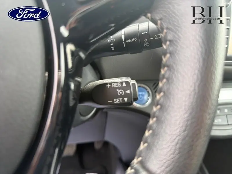 Gros plan sur le volant cuir noir et la commande de régulateur de vitesse d'une Toyota Yaris 116h blanche, vue intérieure côté gauche.