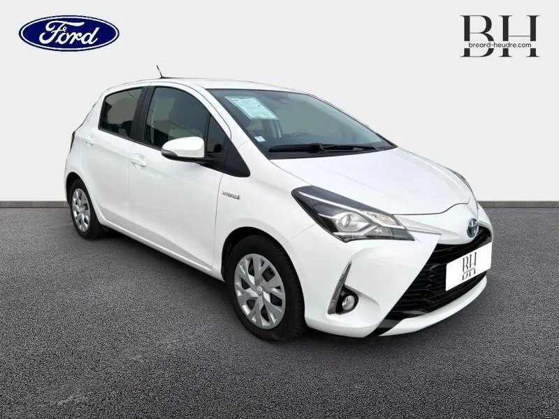 Vue 3/4 avant d'une Toyota Yaris hybride blanche avec enjoliveurs et phares halogènes visibles sur fond neutre.