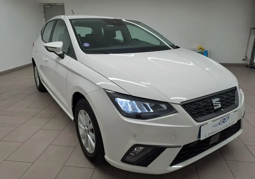 SEAT Ibiza blanc candy vue 3/4 avant droit, phares allumés et calandre noire avec logo central.