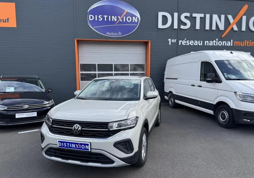 Volkswagen T-Cross gris Ascot en 3/4 avant droit, avec calandre noire et projecteurs LED, devant un garage Distinxion.