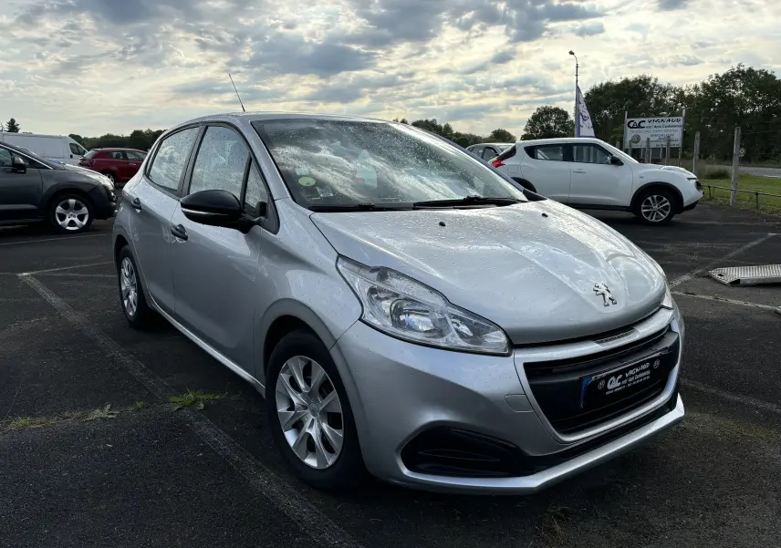 Peugeot 208 gris argent vue 3/4 avant droit sous la pluie sur un parking extérieur avec ciel nuageux.