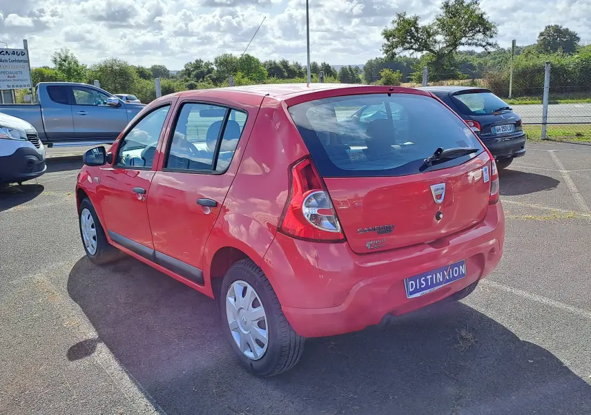 Vue 3/4 arrière droite d'une Dacia Sandero rouge de 2010 avec plaques et badges visibles sur un parking extérieur.