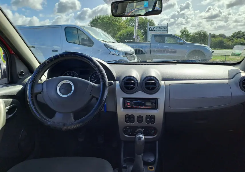 Vue intérieure du tableau de bord et volant noir d'une Dacia Sandero rouge de 2010 avec boîte manuelle et autoradio intégré.