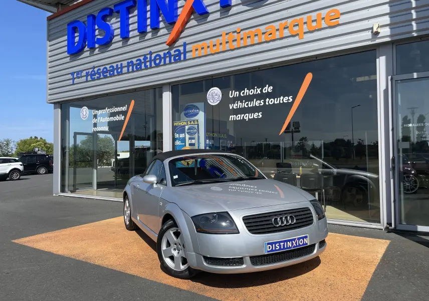 Audi TT Roadster gris argent vue 3/4 avant droit, capote noire, stationné devant un showroom Distinxion.