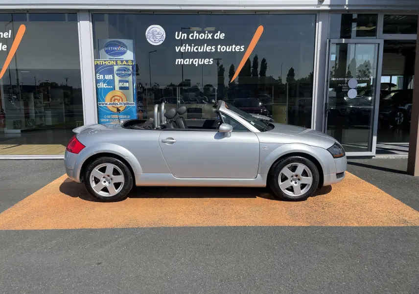 Profil côté gauche d'un Audi TT Roadster gris argent 2002 décapoté, avec jantes 5 branches et intérieur noir visible.